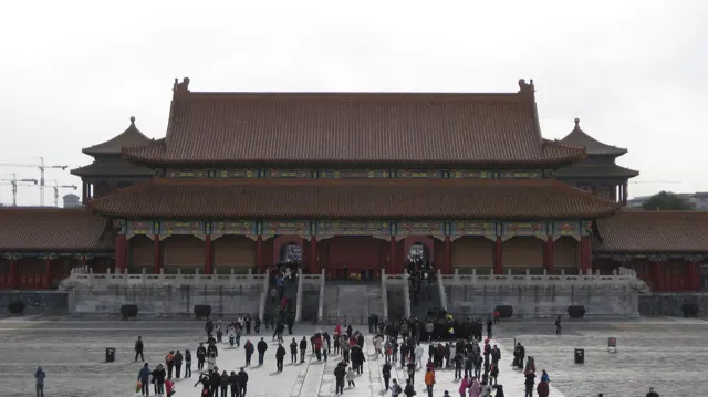 A large red gateway with layered tiled roofs faces visitors crossing the stone-paved space in front of it.