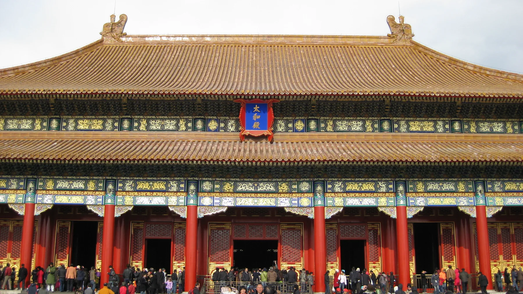 Golden roof tiles and richly painted horizontal beams rise above red columns and busy open entrances.