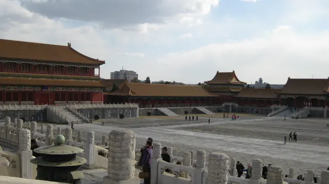 A view from a stone terrace takes in a broad courtyard and long red buildings, with visitors beside the balustrade.