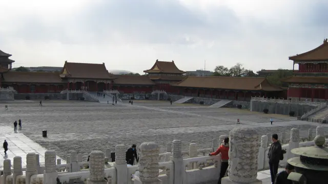 Visitors stand by a carved stone railing overlooking a large courtyard bordered by red walls and tiled roofs.