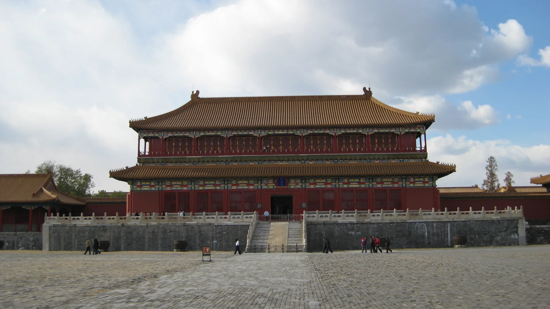 A two-storey red building with a broad tiled roof stands beyond an open paved square, beneath a partly cloudy sky.