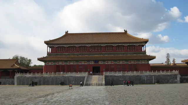 A two-storey red building with a broad tiled roof stands beyond an open paved square, beneath a partly cloudy sky.