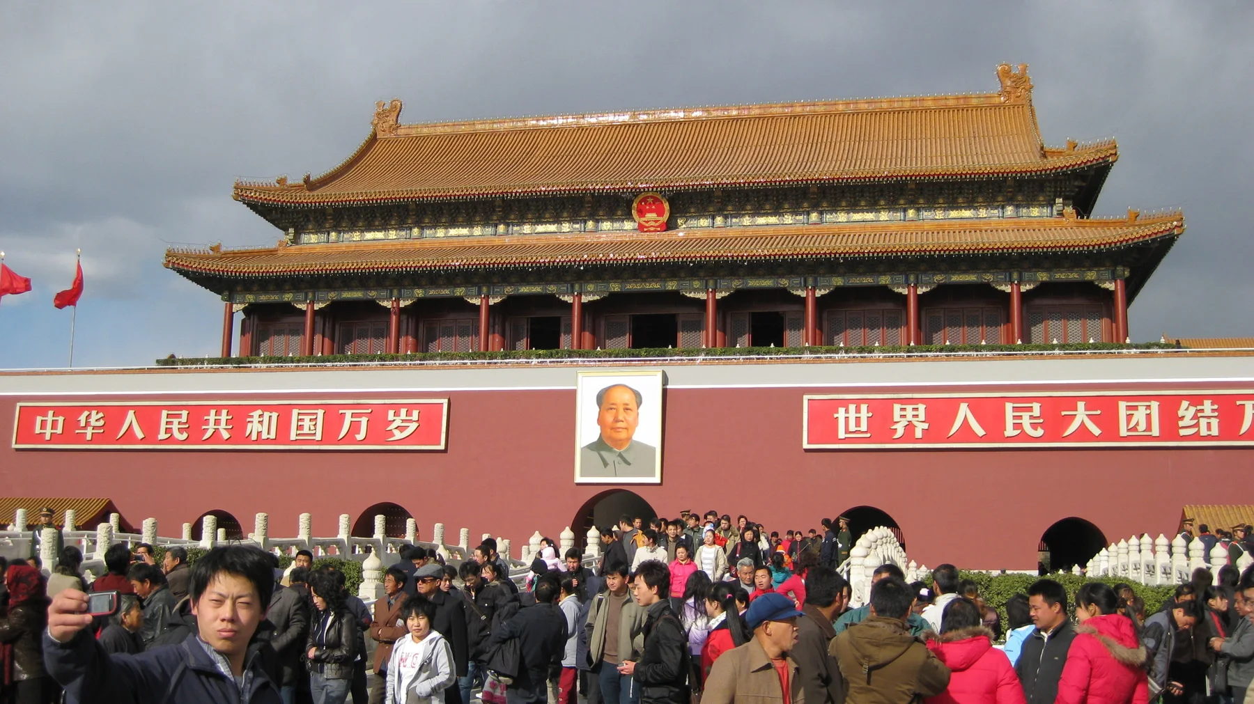 A crowd fills the foreground before a monumental red gate bearing a large portrait, white-lettered signs and a red emblem beneath layered golden roofs.