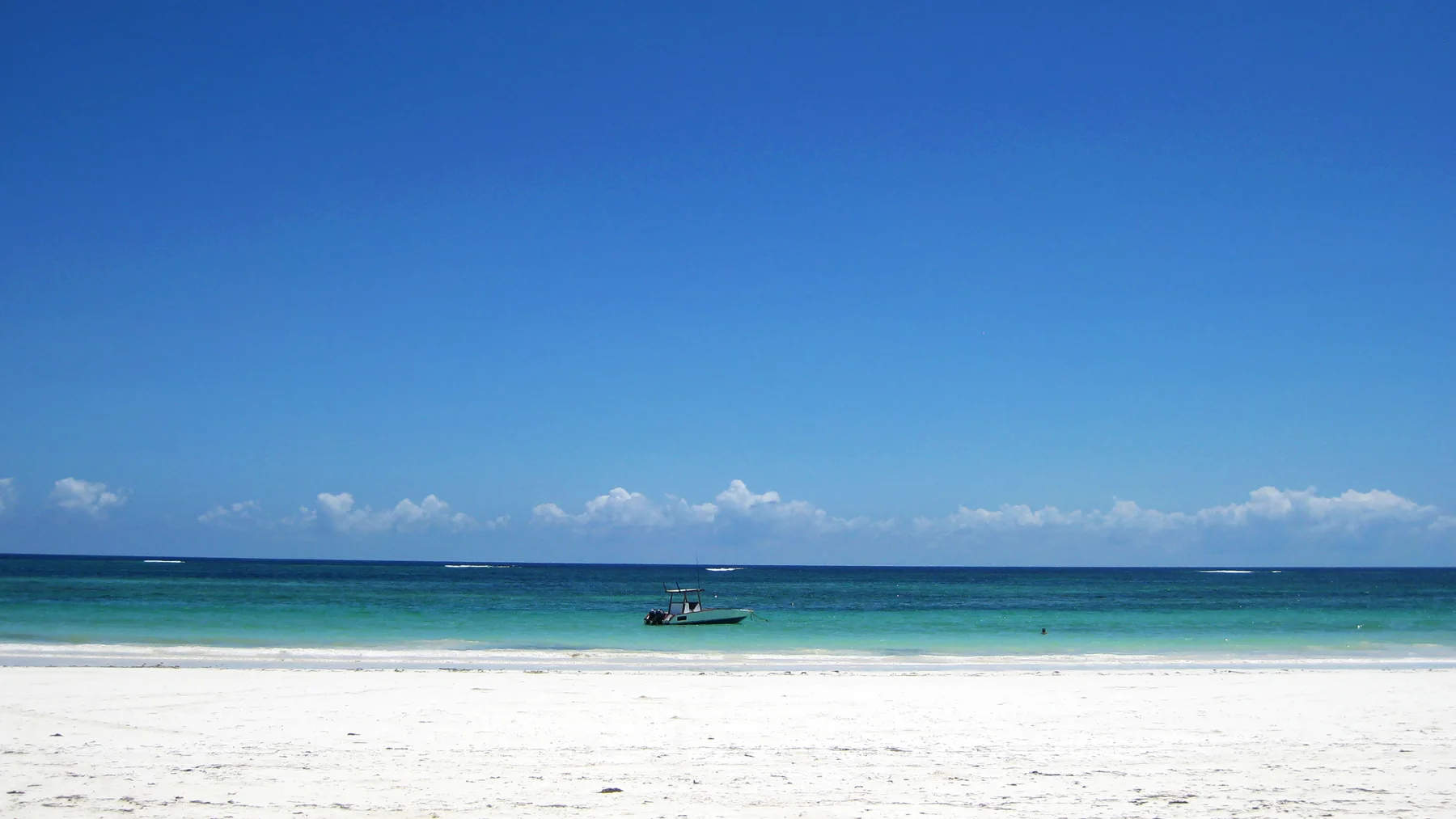 A broad white-sand beach meets turquoise water beneath a clear blue sky, with a small boat offshore.