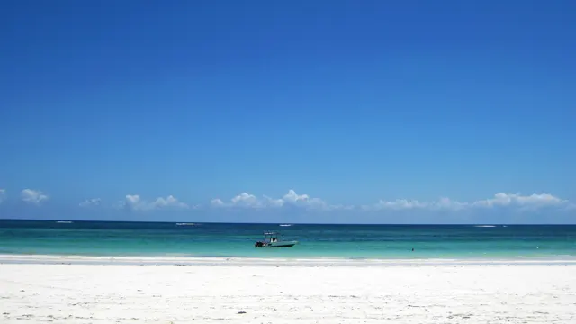 A broad white-sand beach meets turquoise water beneath a clear blue sky, with a small boat offshore.