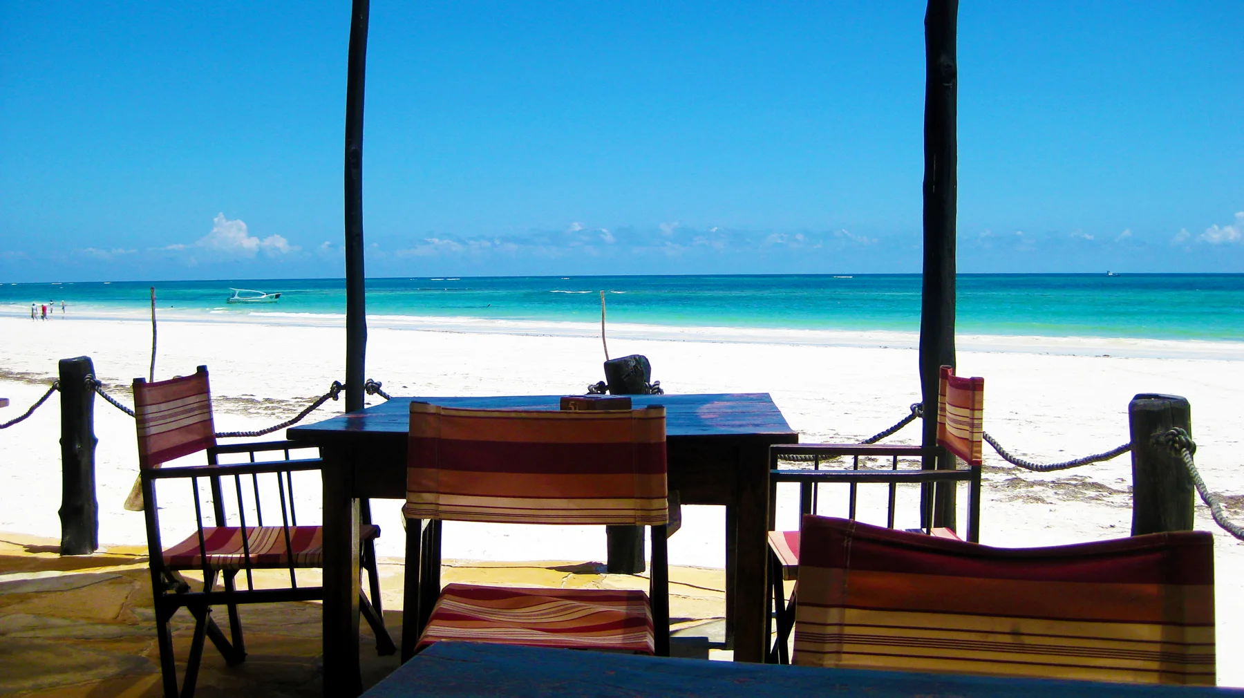 Striped chairs and a table sit in shade beside a rope barrier, looking out across white sand towards turquoise water.