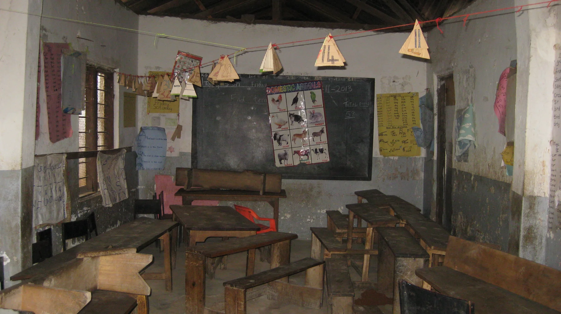 Wooden desks and benches fill a classroom with a blackboard, wall displays and paper shapes suspended overhead.