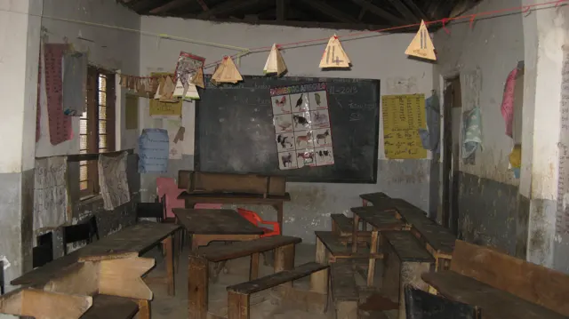 Wooden desks and benches fill a classroom with a blackboard, wall displays and paper shapes suspended overhead.