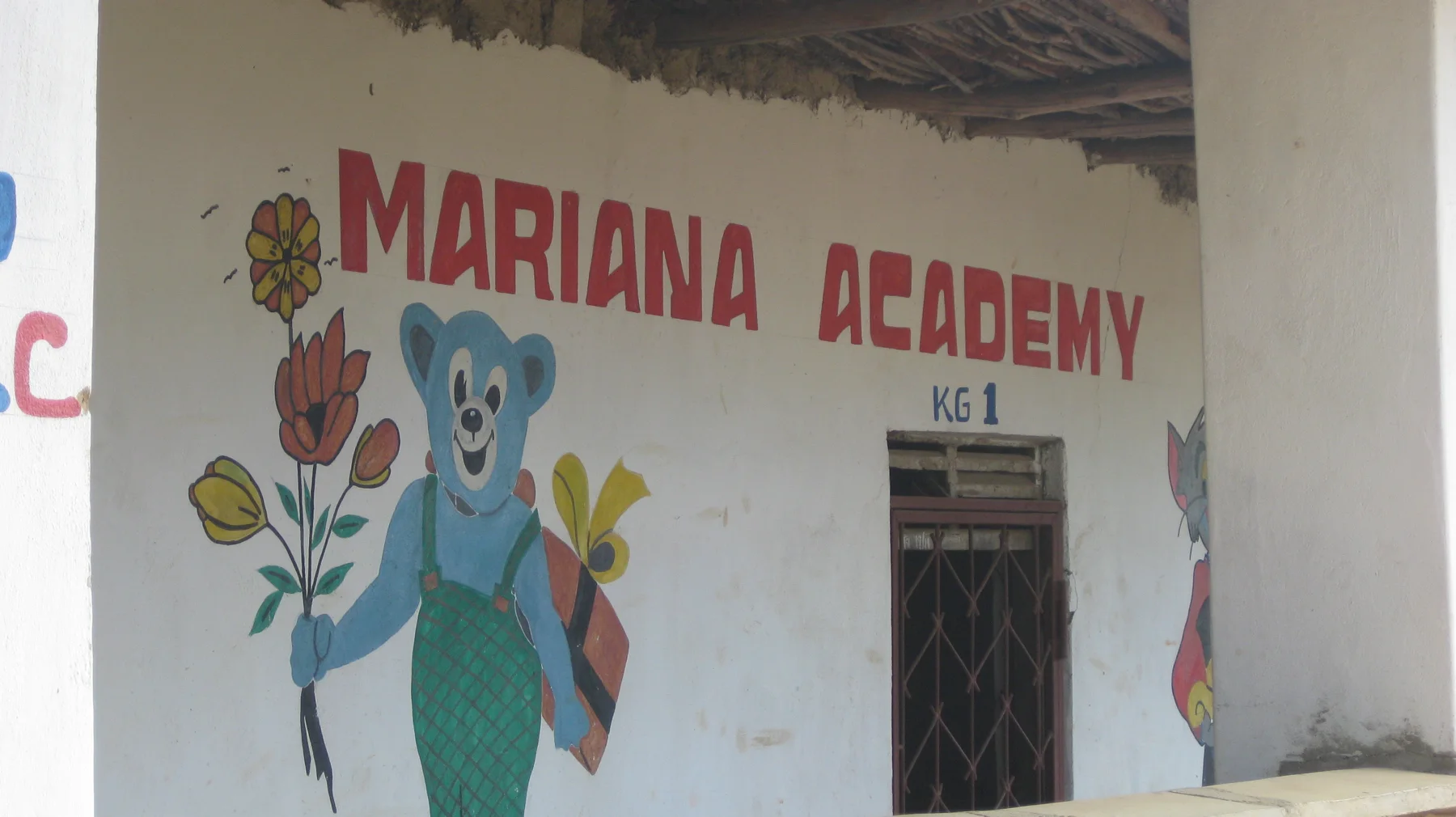 Red school lettering and a painted blue character holding flowers decorate a white wall beside a doorway marked KG 1.