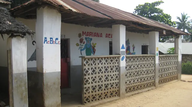 A low school building has a shaded veranda behind patterned concrete blocks, with painted pictures on the wall and sandy ground in front.