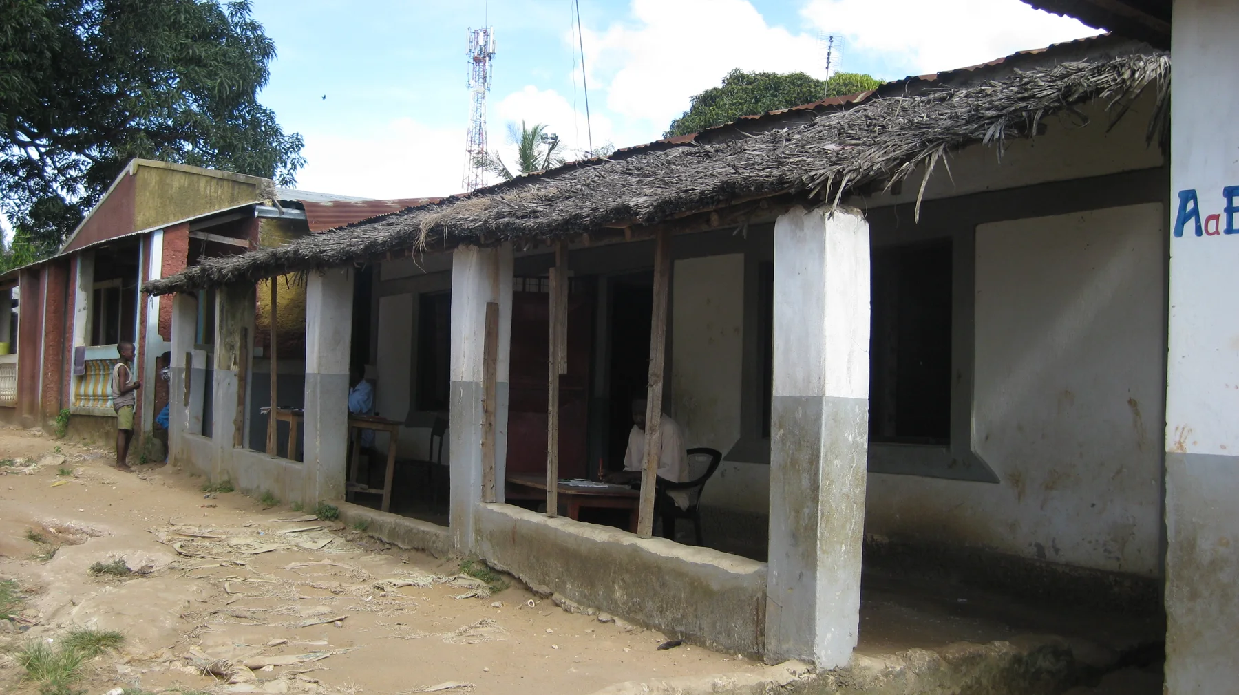A row of low buildings with shaded doorways and white veranda pillars runs beside an unpaved courtyard.