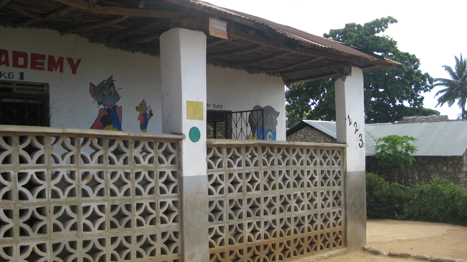 Decorative concrete blocks line a school veranda beneath white pillars and a corrugated roof, with colourful wall paintings behind.