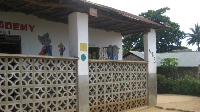 Decorative concrete blocks line a school veranda beneath white pillars and a corrugated roof, with colourful wall paintings behind.