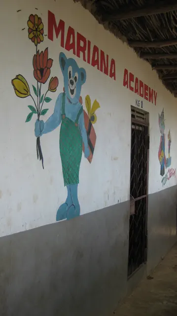 An angled portrait view follows a white school wall with red lettering and a large blue painted character beside a doorway.