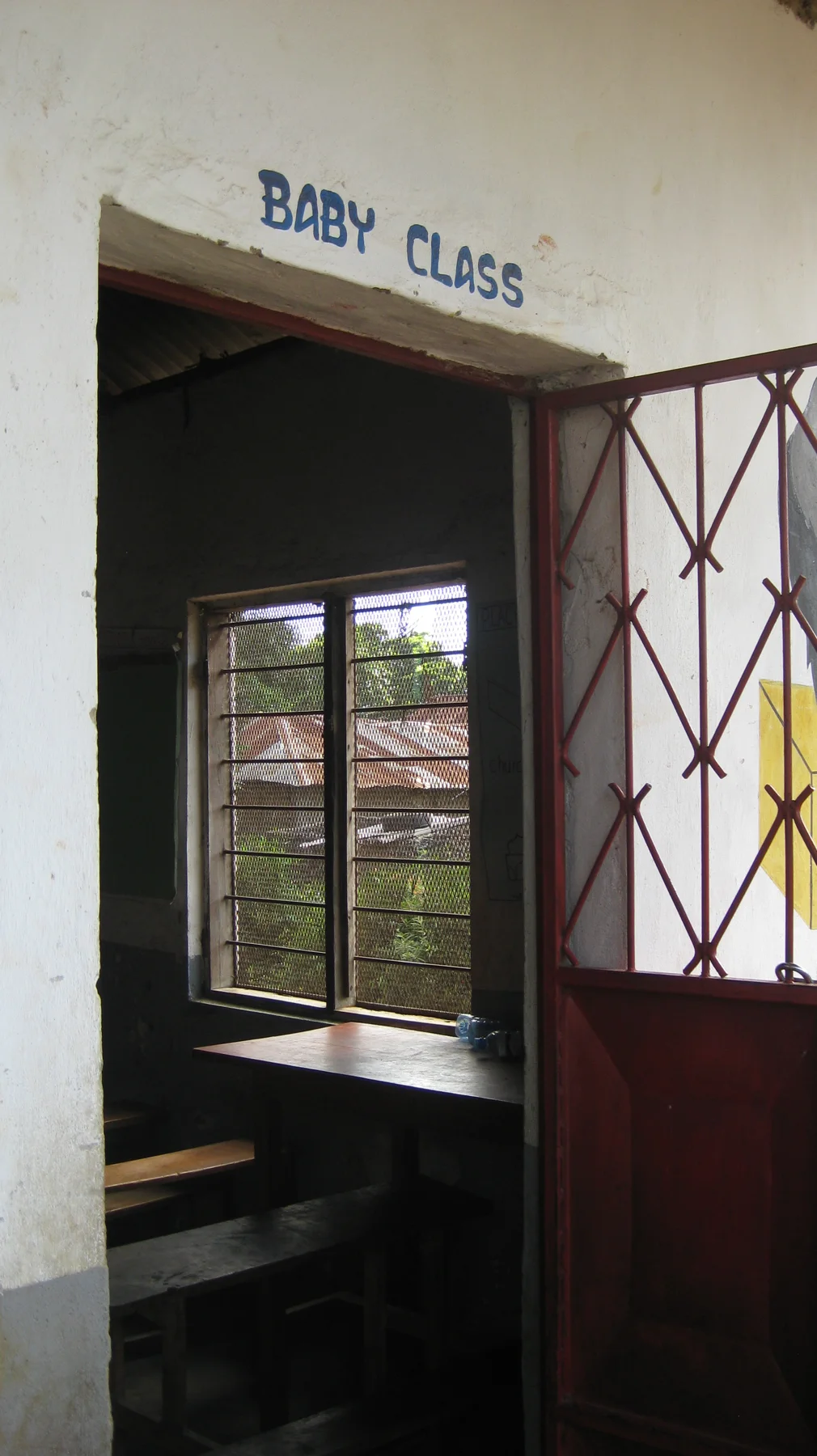 An open red metal door leads into a classroom beneath a sign reading Baby Class, with a window and desks visible inside.