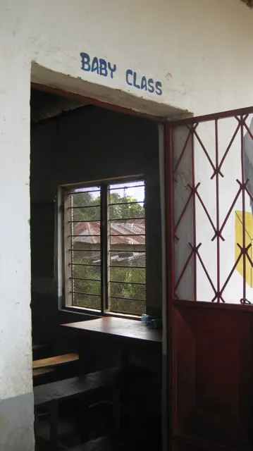 An open red metal door leads into a classroom beneath a sign reading Baby Class, with a window and desks visible inside.