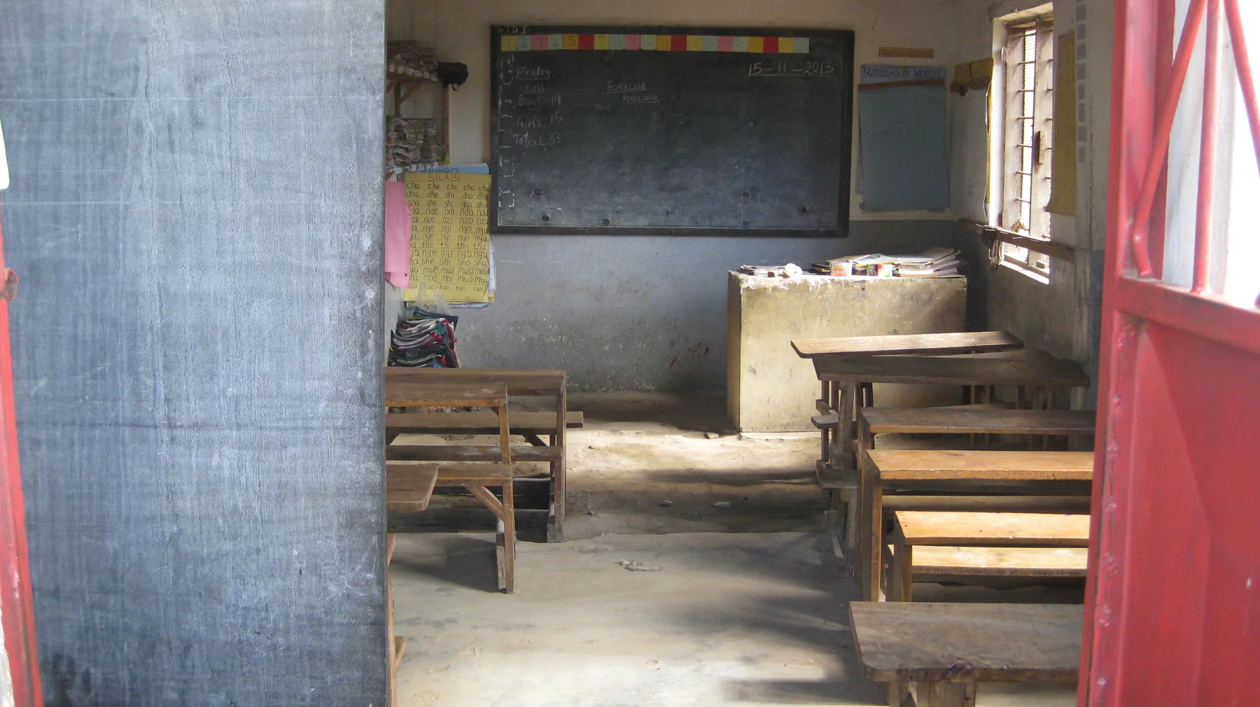 A view through a red doorway shows wooden benches, a blackboard and a small raised surface beneath a bright classroom window.