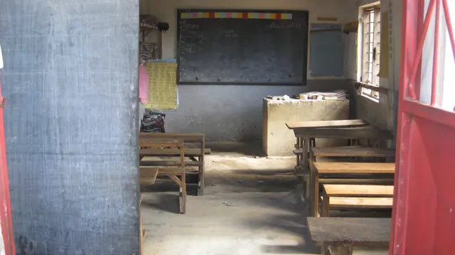 A view through a red doorway shows wooden benches, a blackboard and a small raised surface beneath a bright classroom window.