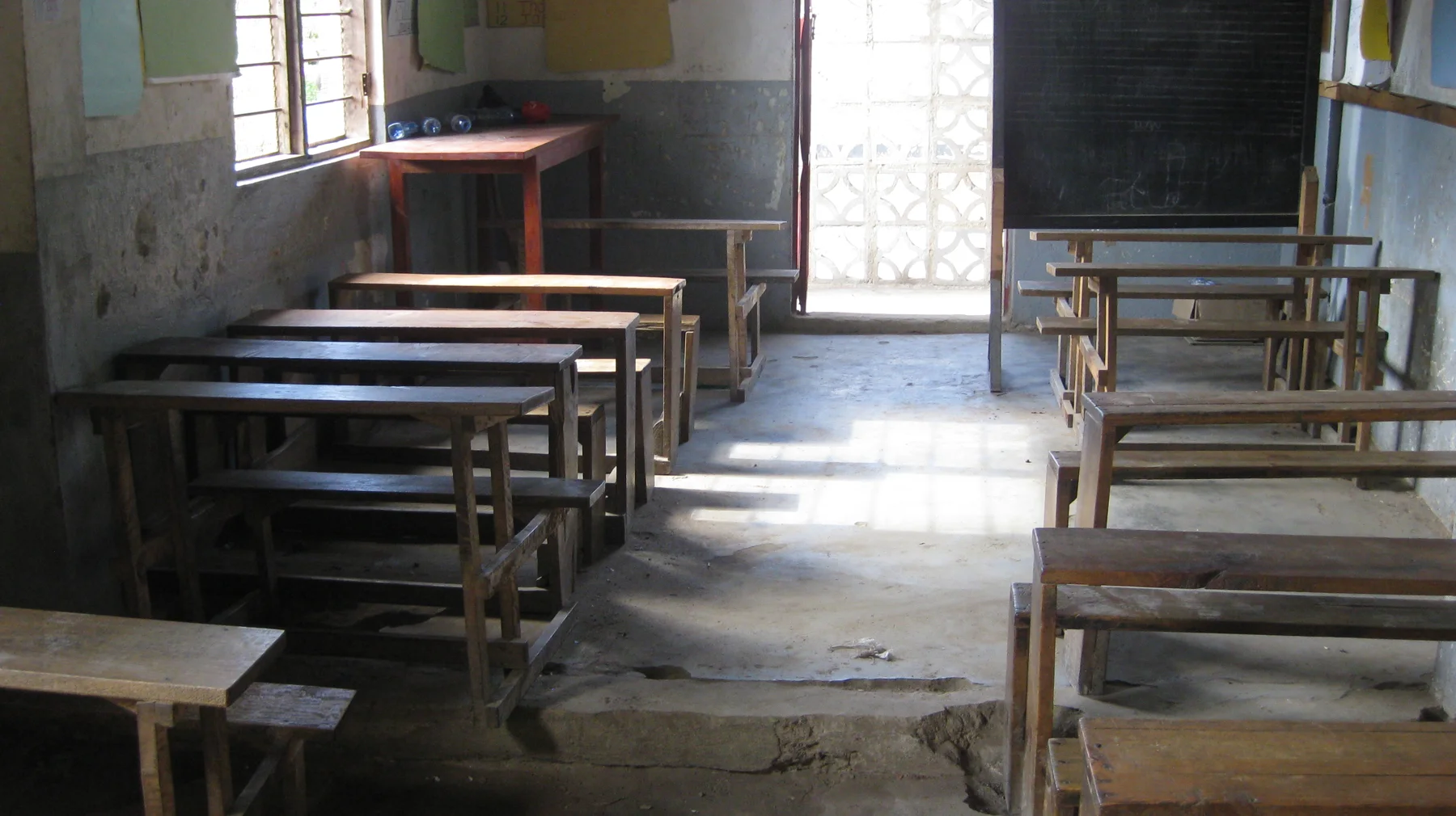 Wooden desks and benches stand in rows inside a classroom, with daylight entering through a window and a doorway beyond.