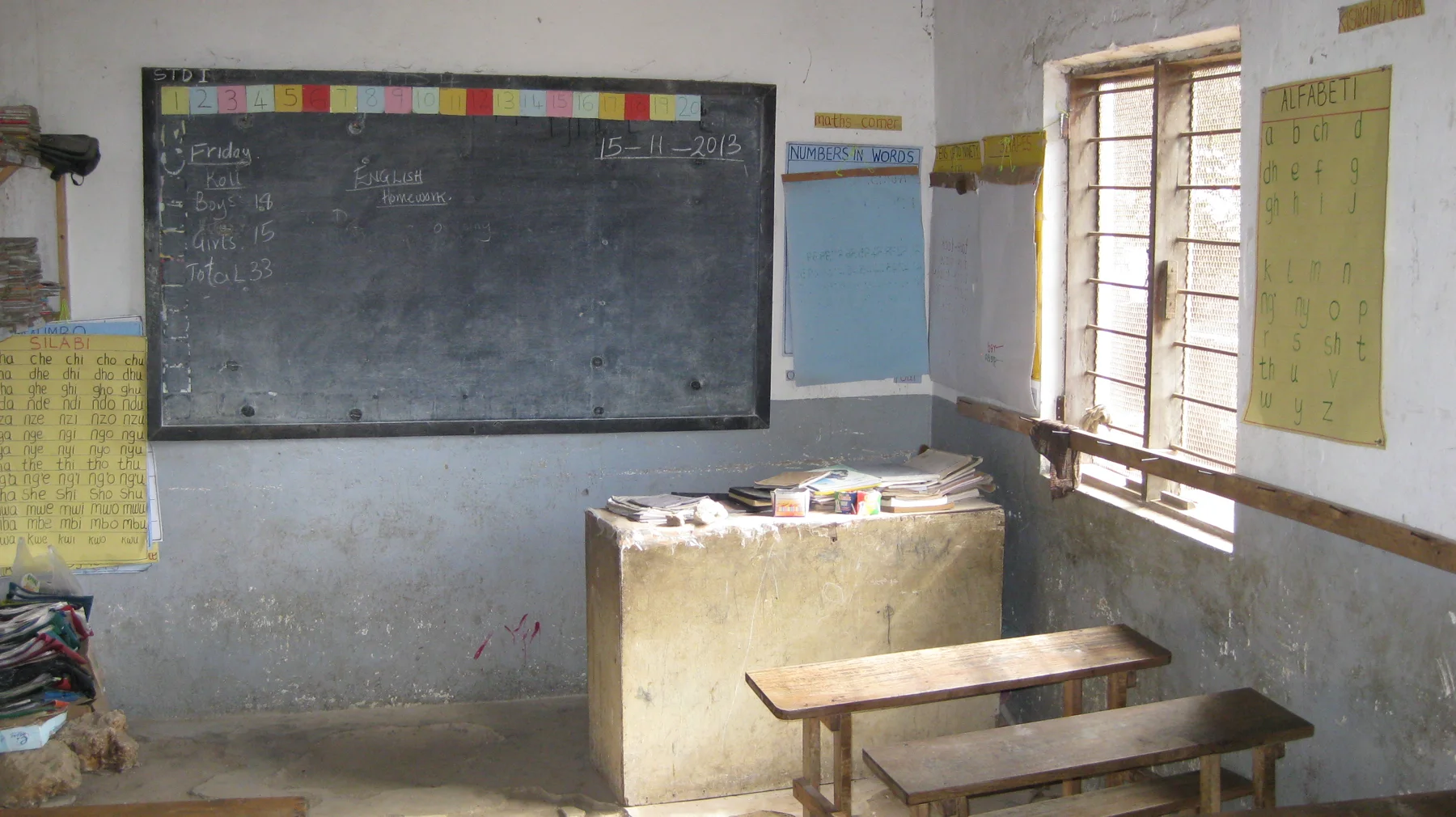 A dark classroom blackboard hangs beside a bright window and paper wall charts, above a small raised surface and wooden bench.