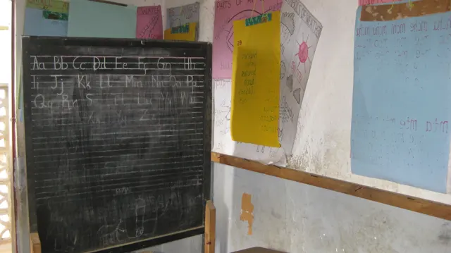 Handwritten letters and ruled lines cover a classroom blackboard beside colourful paper charts on the wall.