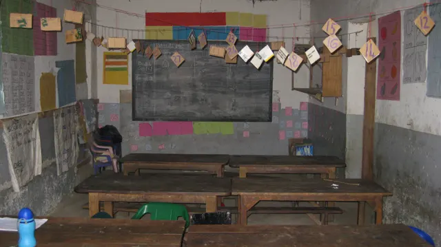 Strings of paper cards hang above wooden desks in a classroom with a central blackboard and colourful wall displays.