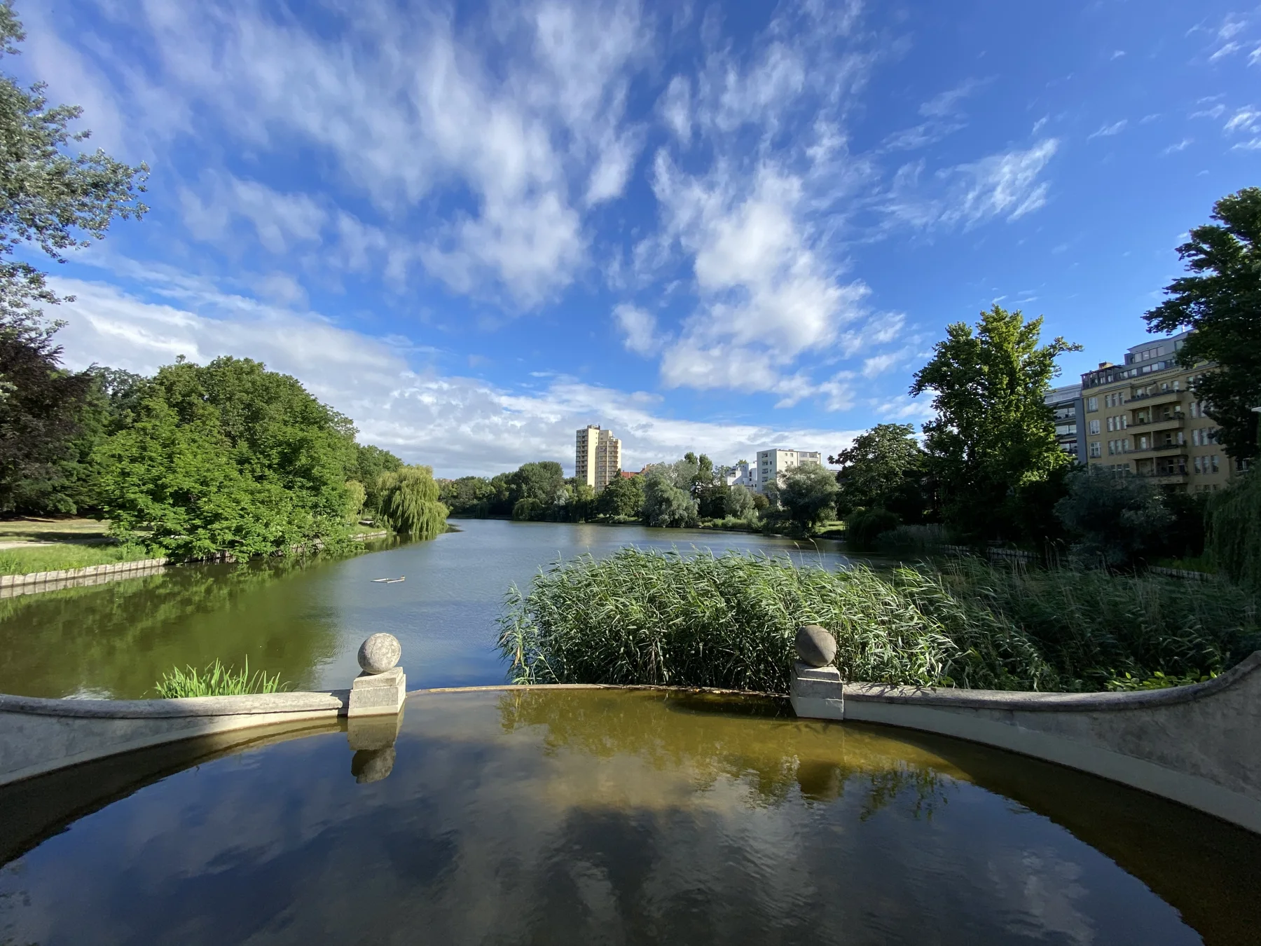 A view over a shallow foreground pool towards a tree-lined lake, with buildings along the far shore beneath a blue sky streaked with clouds.