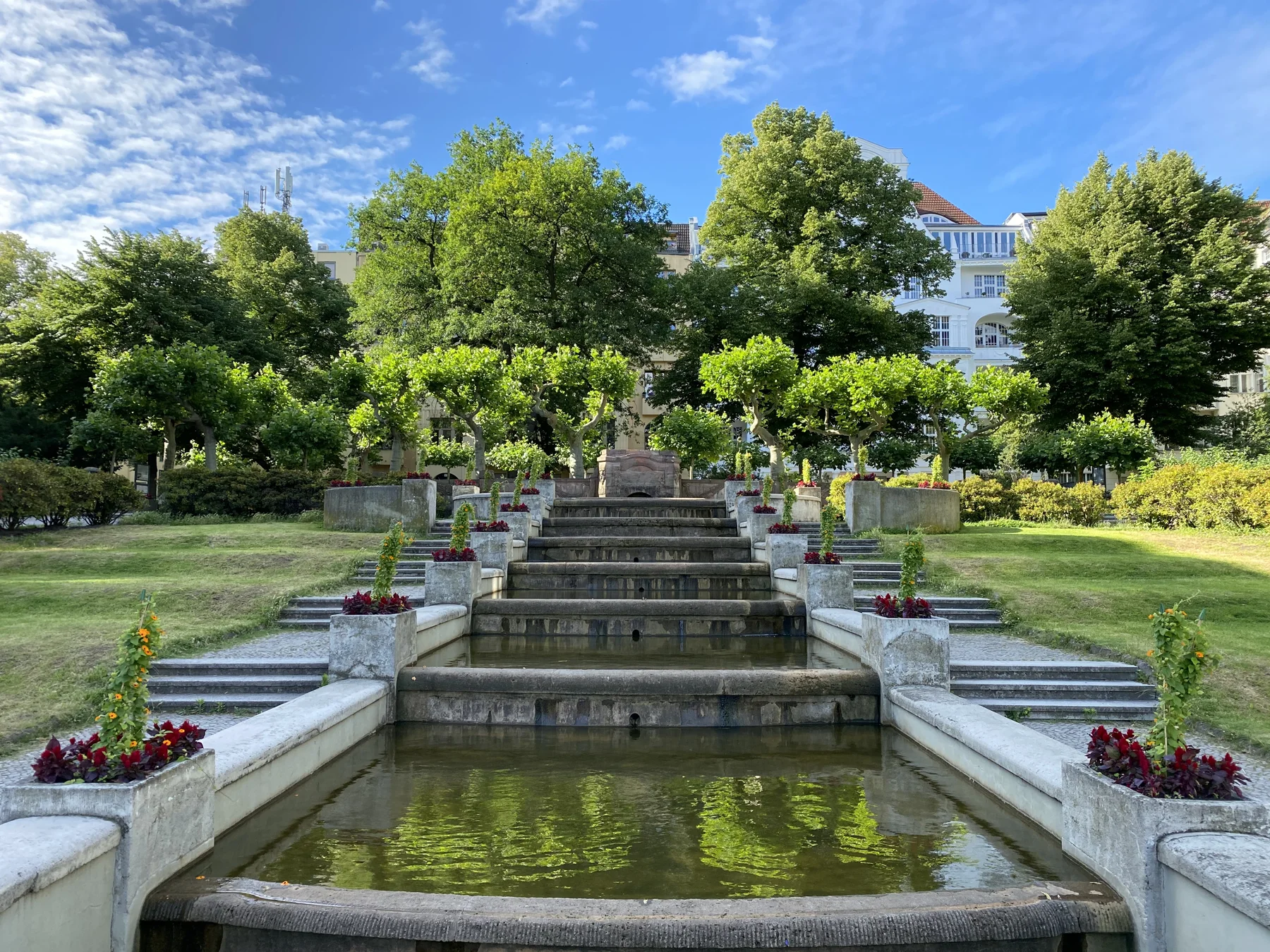 A series of rectangular pools descends between pale walls and planted urns, with steps, lawns and trees beyond.