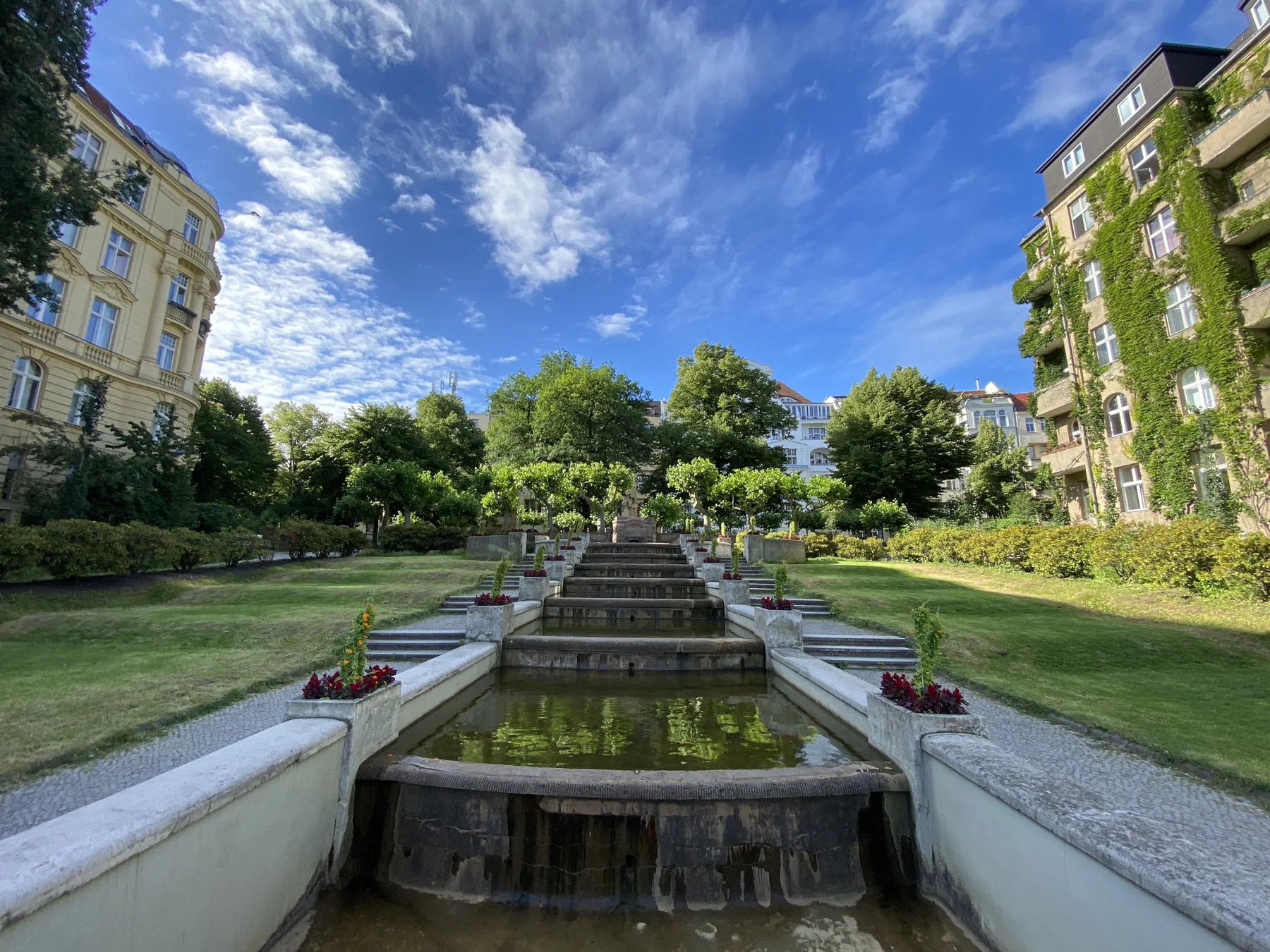 A wide view up a stepped cascade, framed by lawns and apartment buildings beneath a blue sky with wispy clouds.