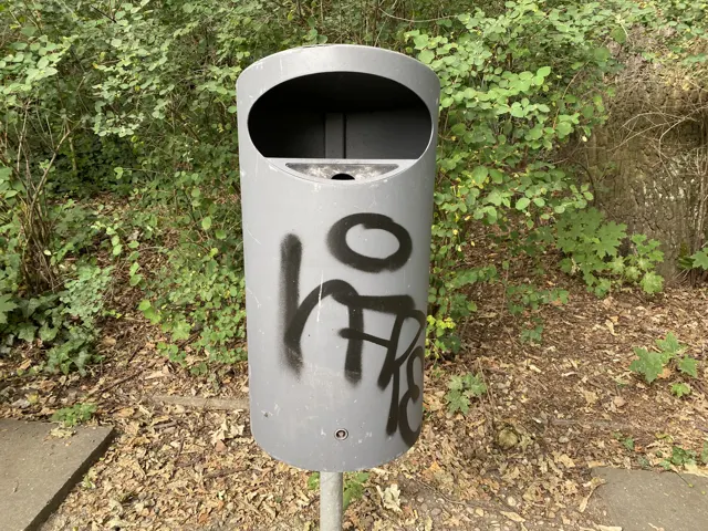 A grey cylindrical litter bin marked with black graffiti stands beside a path, backed by dense green shrubs.