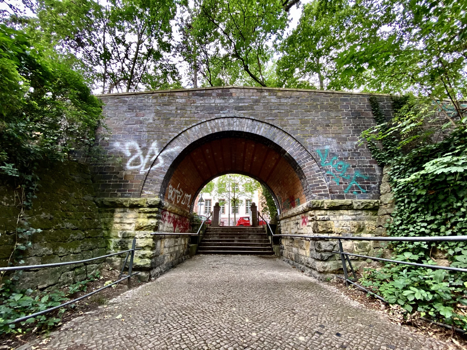 A path leads through a brick arch marked with graffiti, with stonework at its base and leafy trees around it.