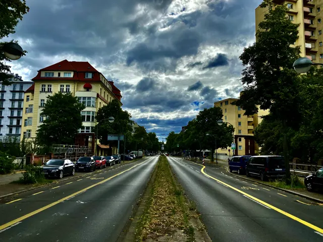 Two roadways run beside a planted central strip, with parked cars and apartment buildings under a dramatic cloud-filled sky.