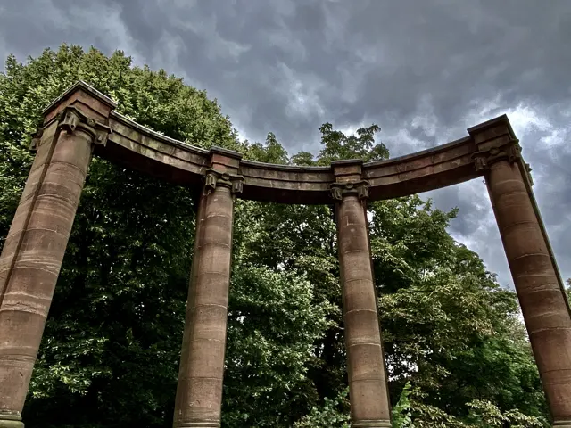 A curved stone structure supported by tall columns rises against grey clouds, with green treetops behind it.