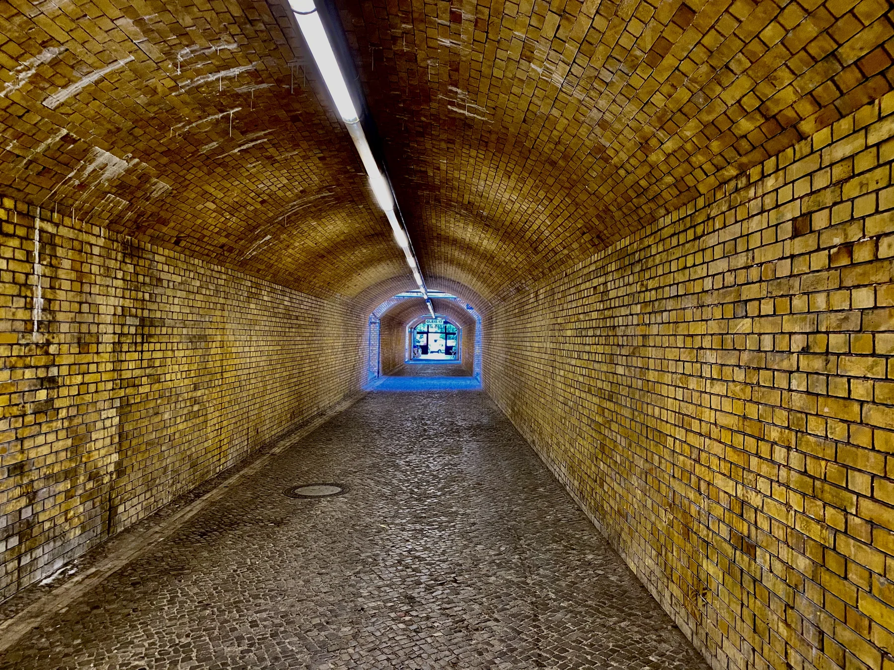 A long vaulted passage lined with yellow-brown bricks leads towards a distant opening, beneath a narrow ceiling light.