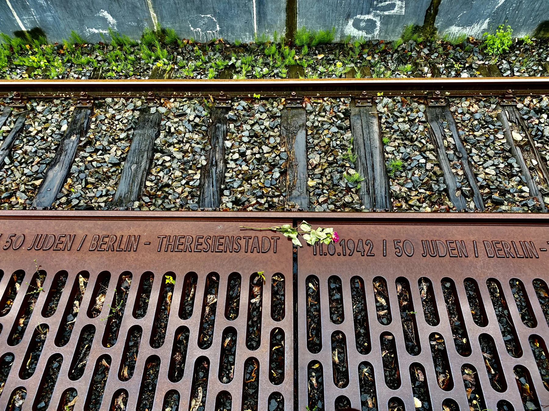An inscribed metal platform edge lies beside railway sleepers and gravel, with small flowers resting near the lettering.