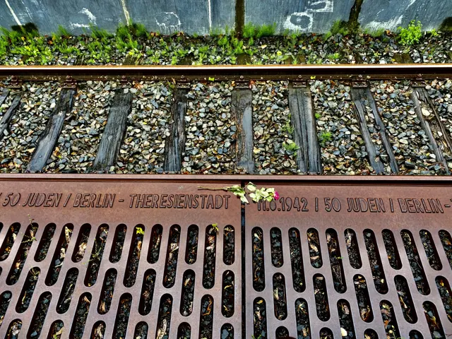 An inscribed metal platform edge lies beside railway sleepers and gravel, with small flowers resting near the lettering.