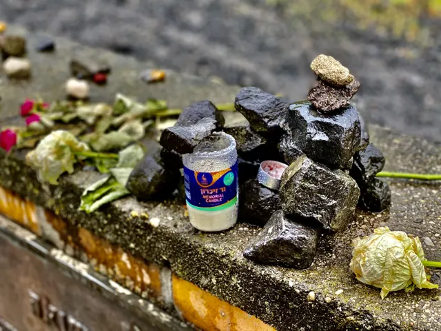 A memorial candle stands beside stacked stones and flowers on a ledge, with a pale flower in the foreground.