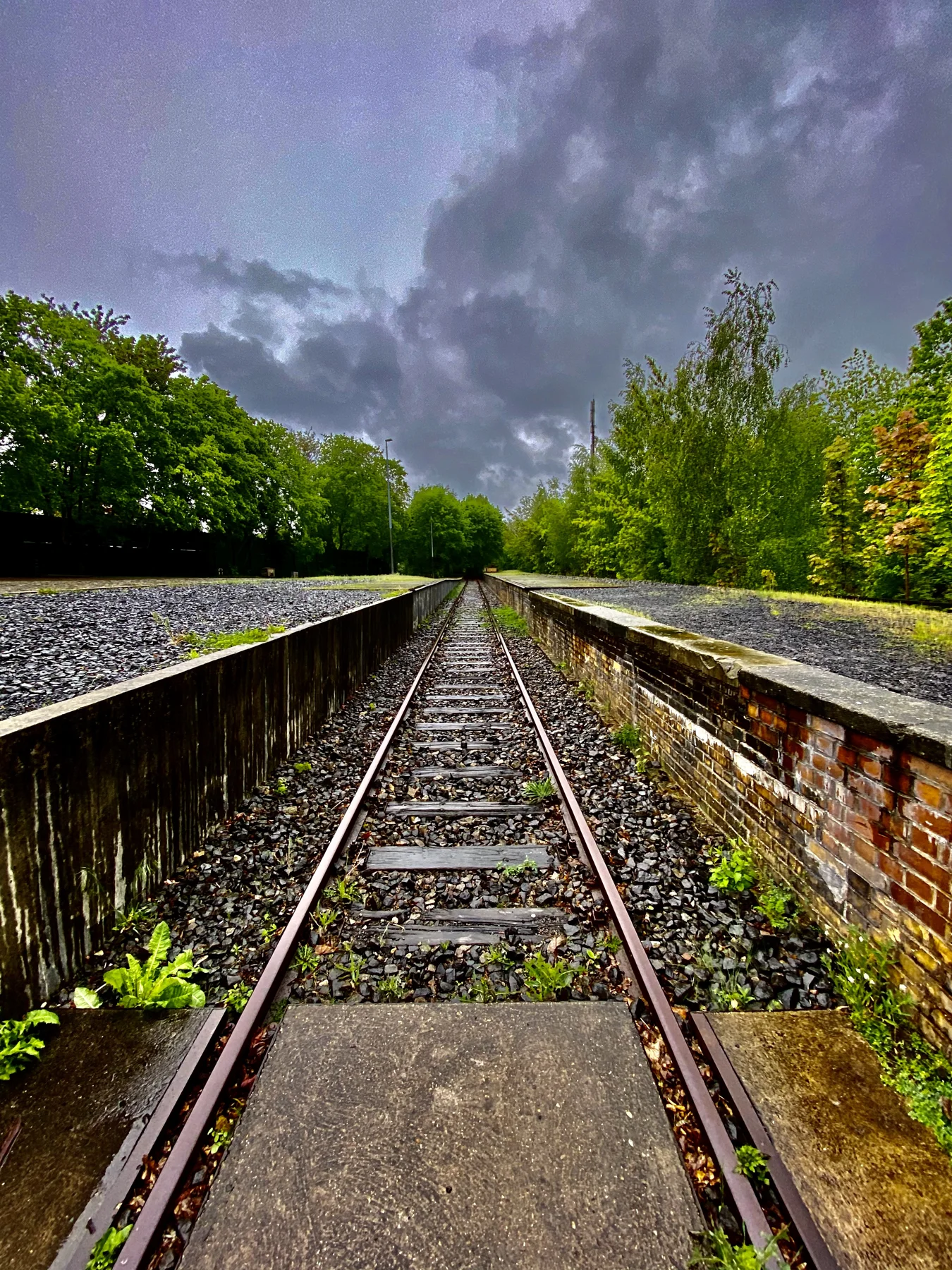 A railway track runs between raised platform edges towards green trees beneath dark clouds, seen in a portrait composition.