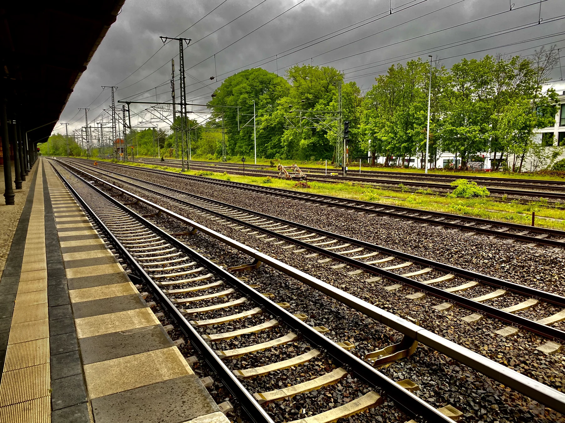 Parallel railway tracks and overhead wires extend beside a covered station platform, with trees and buildings across the rails.