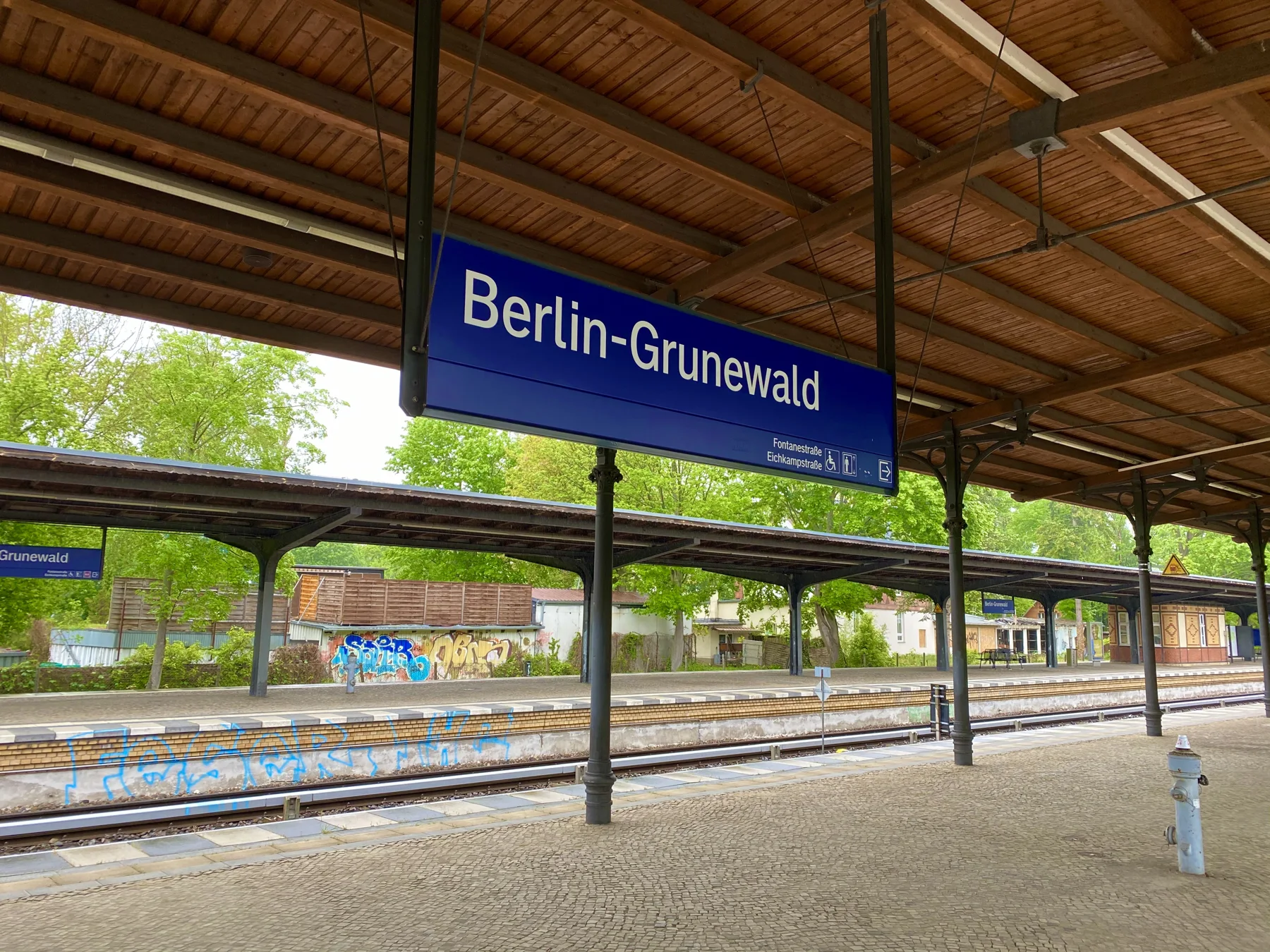 A blue Berlin-Grunewald station sign hangs beneath a wooden platform roof, with tracks and another platform beyond.