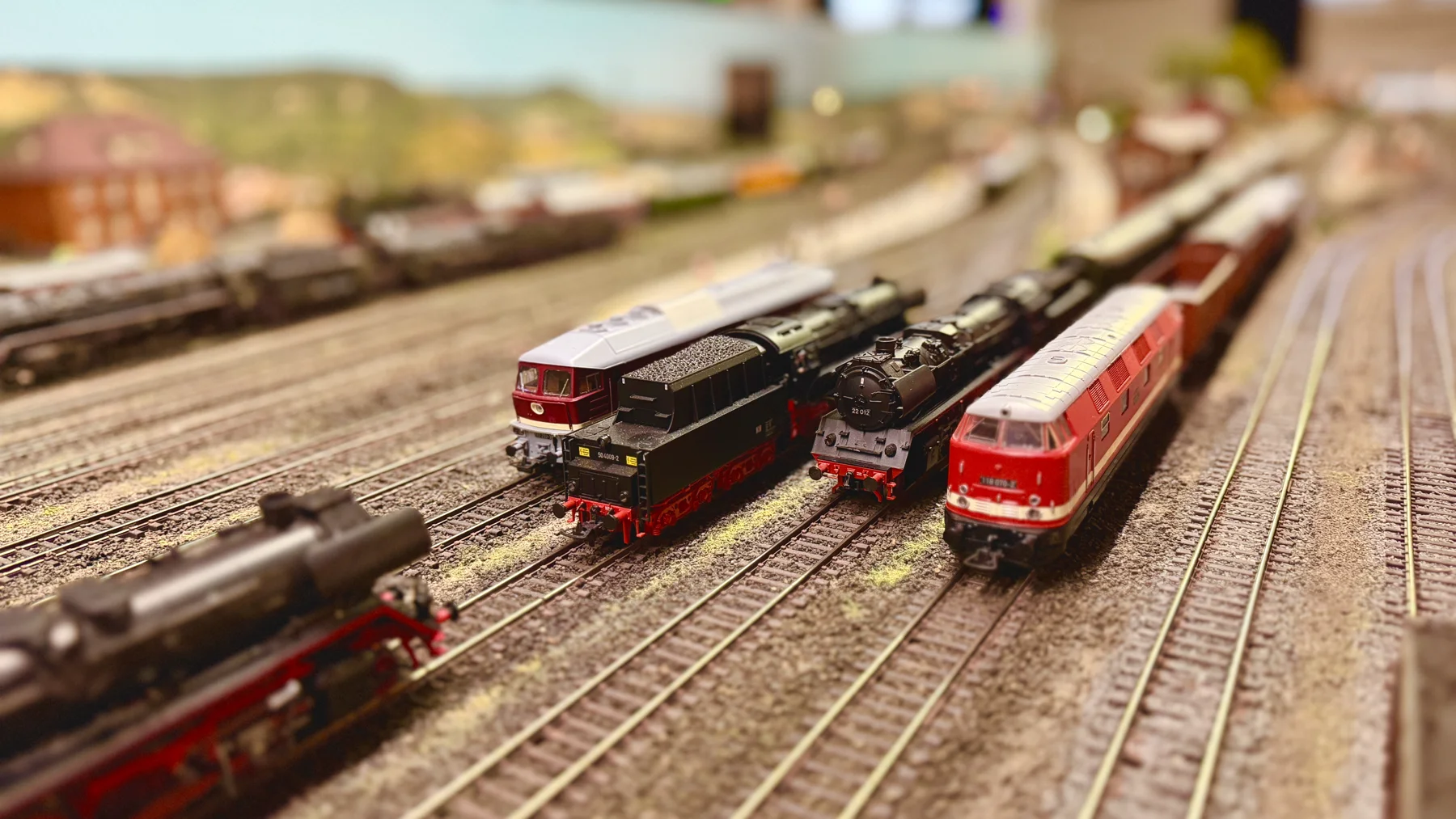 Red and dark model locomotives stand side by side on parallel railway tracks.