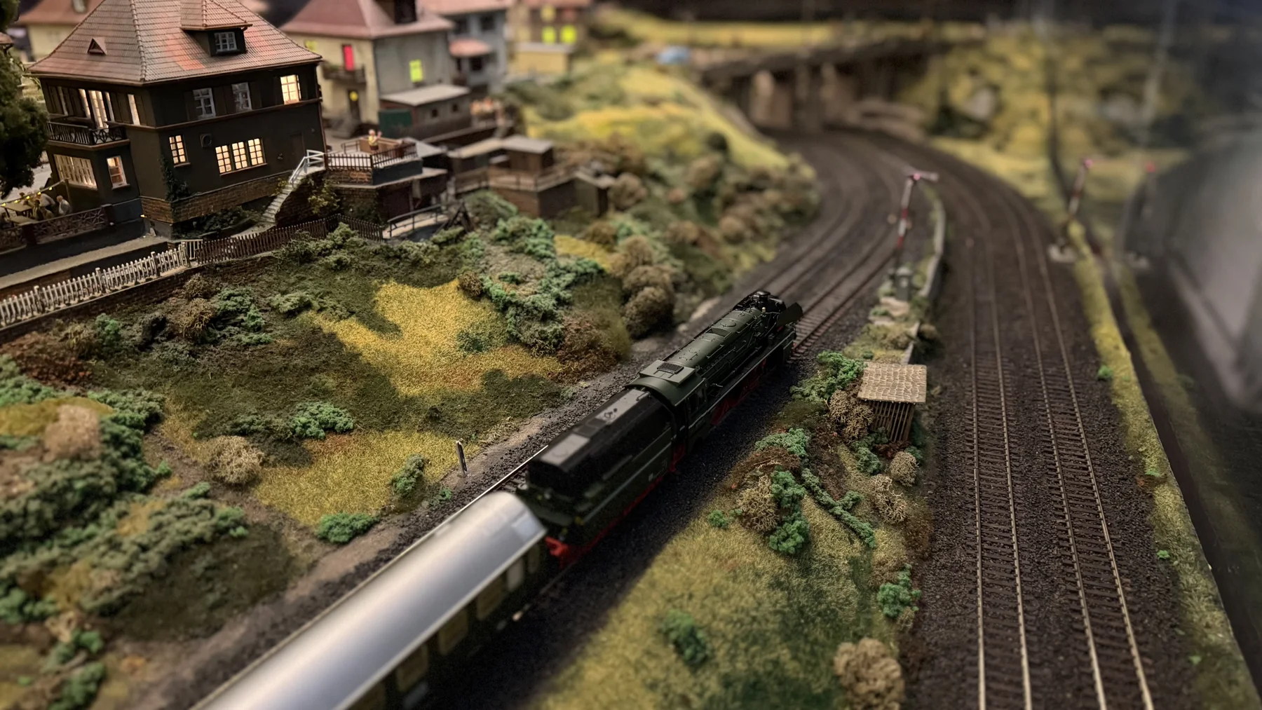 A model train follows a curved track past a small house and miniature trees.