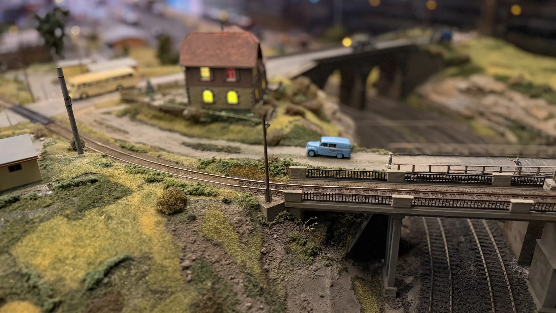 A miniature road bridge crosses railway tracks beside a small house and a blue car.