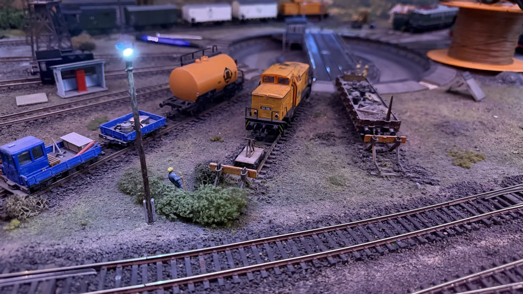 Small blue vehicles, an orange tank and freight wagons fill a model railway yard.