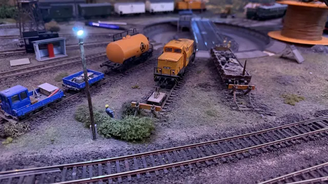 Small blue vehicles, an orange tank and freight wagons fill a model railway yard.