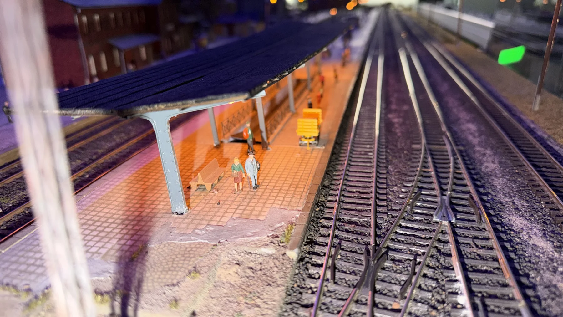 An orange-lit model platform with tiny figures stands beside empty railway tracks.