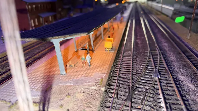An orange-lit model platform with tiny figures stands beside empty railway tracks.