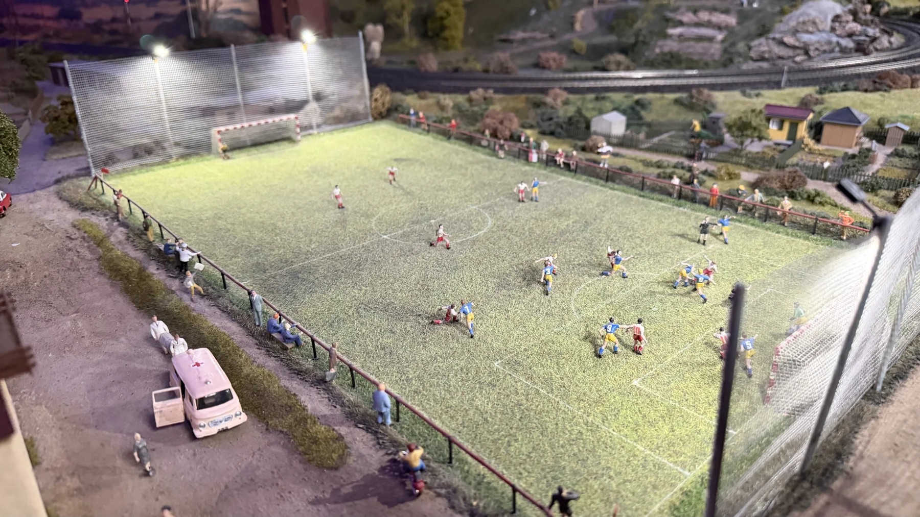 Tiny players fill a miniature football pitch under bright floodlights beside a railway.
