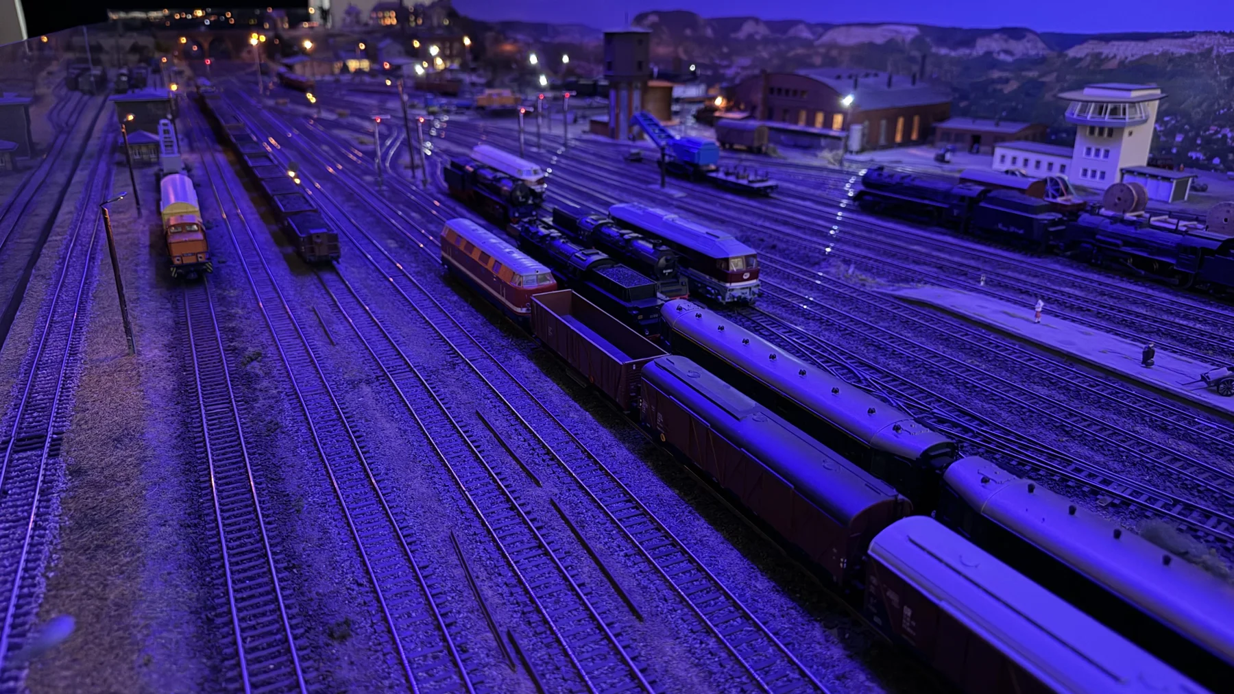 A wide model railway yard with several trains and buildings is washed in blue light.