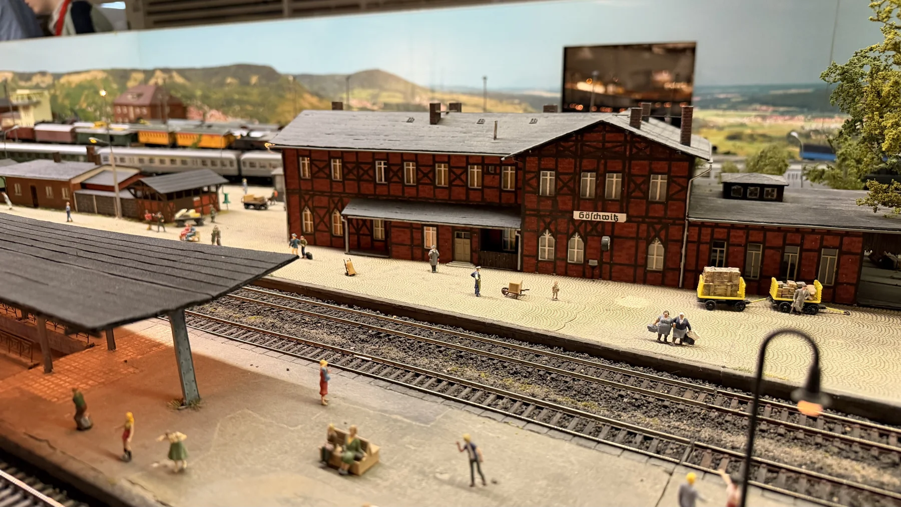 Tiny figures stand outside a large brick station building behind a covered platform.